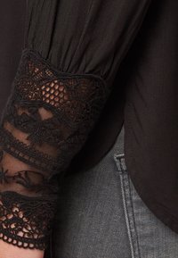 Schwarze Bluse mit gerafftem Stoff am Ärmel, Spitzenbesatz mit floralen Mustern, transparenter Textur und filigranen Details am Bündchen.