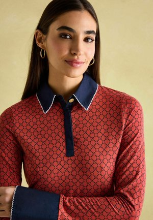 Red patterned polo shirt with a navy collar and button, featuring white scalloped detailing on the collar and cuffs. Soft fabric texture.