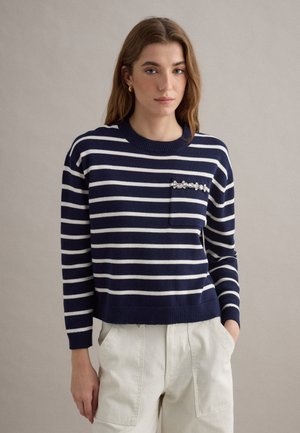 Junge Frau trägt einen marineblauen und weiß gestreiften Pullover mit verziertem Brusttaschen-Detail und cremefarbene Hose, steht vor einfachem Hintergrund.