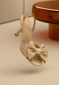 Beige høyhælte sandaler med teksturert stoff, åpen tå og stor sløyfedetalj foran. Ankelrem med gullfarget hardware.