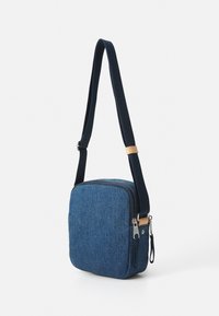Blå denim crossover-veske med firkantet form, som har glidelås, justerbar stropp og skinndetalj på stroppen.