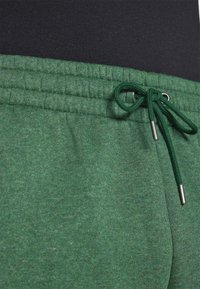 Grønne sweatpants med struktureret stof, elastisk talje og matchende snøre med metalspidser. Sort talje synlig ovenfor.