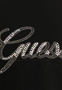 Svart t-shirt med rhinestones som bildar namnet "Guess" med silveraccenter och broderade detaljer.