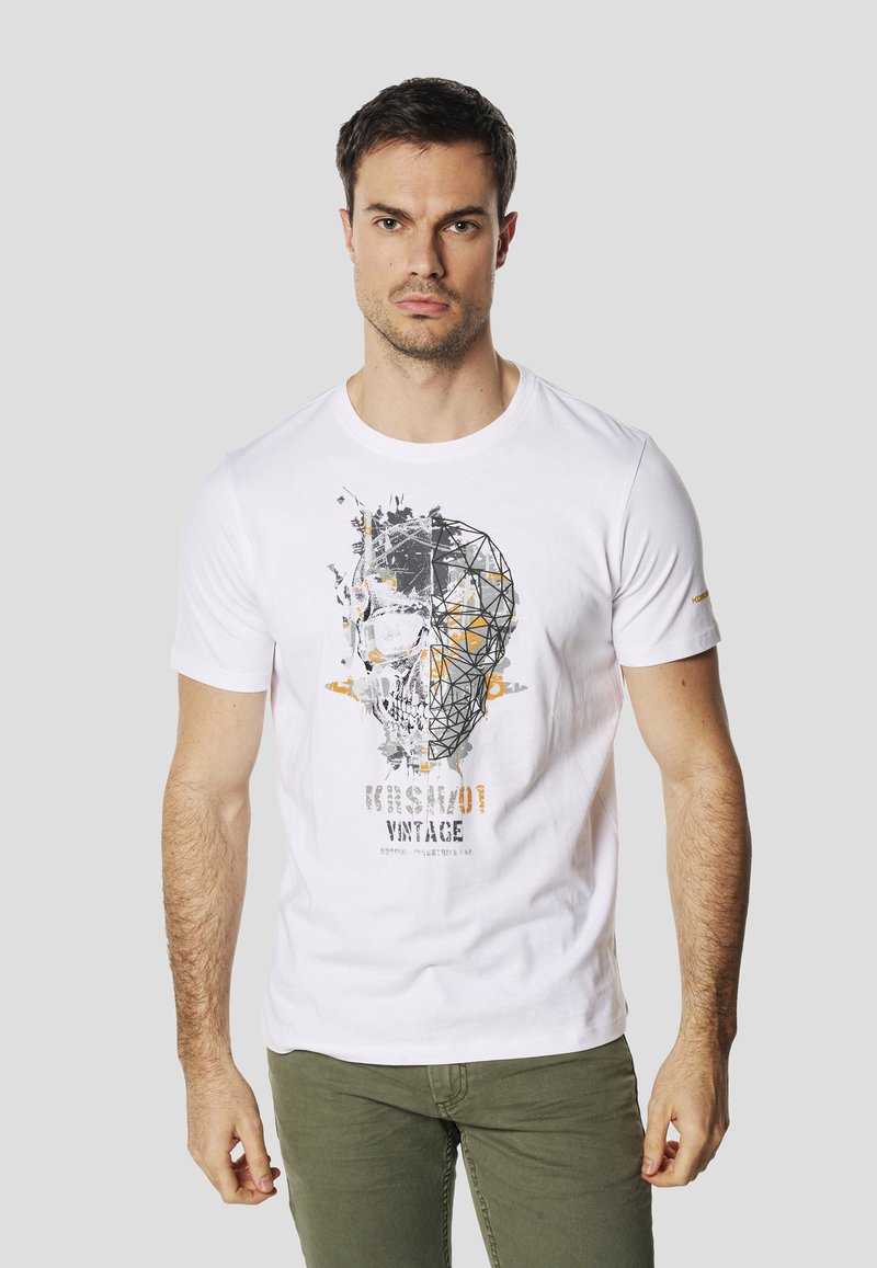 T-shirt en coton blanc avec un motif géométrique de crâne en gris et orange, avec le texte "Kris Scott Vintage" imprimé en dessous. Manches courtes.