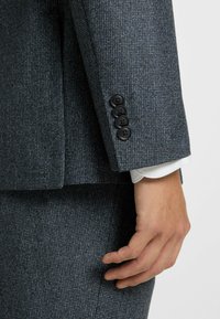 Grijze textuur blazer met notch-lapel, voorzien van vier zwarte knopen op de manchet en zichtbare witte shirtmanchetten eronder.