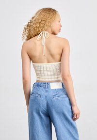 Top halter de canalé color crema claro con cierre de lazo, combinado con pantalones de mezclilla azul de tiro alto que tienen bolsillos traseros y una etiqueta en blanco.