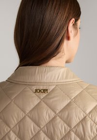 Beige gewatteerde jas met een gestructureerde afwerking, voorzien van een gouden logo "JOOP!" nabij de kraag. De kraag is klassiek en gestructureerd.