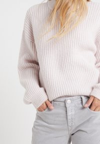 Jasnoróżowy sweter z prążkowanej tkaniny z długimi rękawami, z wysokim dekoltem, w połączeniu z jasnoszarymi spodniami, podkreślonymi subtelnym detalem w postaci guzików.