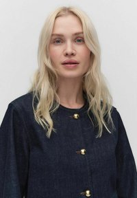 Veste en denim bleu foncé avec un col rond et quatre fermetures à boutons dorés, présentant une texture lisse et des détails de couture minimaux.