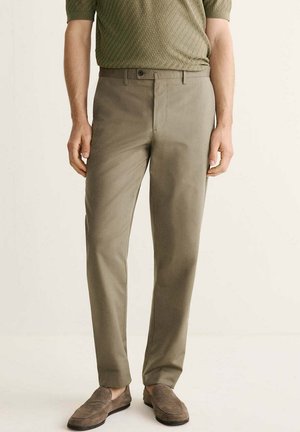 Mann trägt hellbraune Slim-Fit-Hosen, strukturiertes olivgrünes Hemd und braune Wildleder-Loafer und steht vor einem einfarbigen Hintergrund.