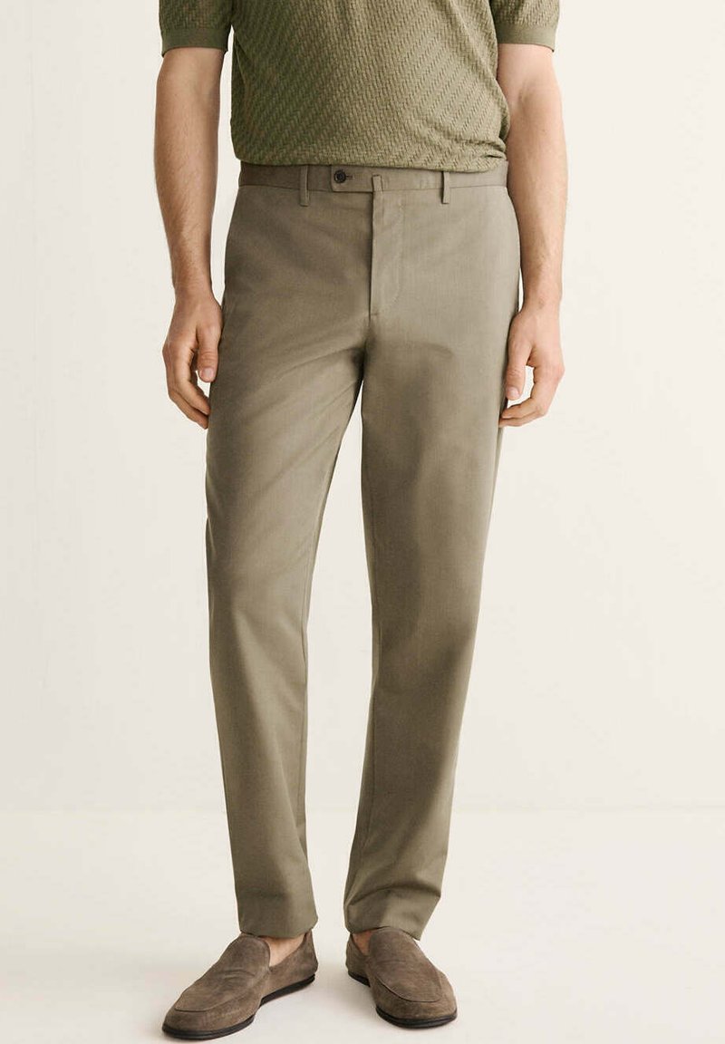 Hombre con pantalones ajustados marrón claro, camisa texturizada verde oliva y mocasines de ante marrones, de pie frente a un fondo liso.