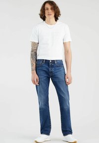 Blauwe spijkerbroek met een rechte snit, gecombineerd met een witte t-shirt met ronde hals voorzien van een subtiel logo. Witte sneakers maken de look compleet.