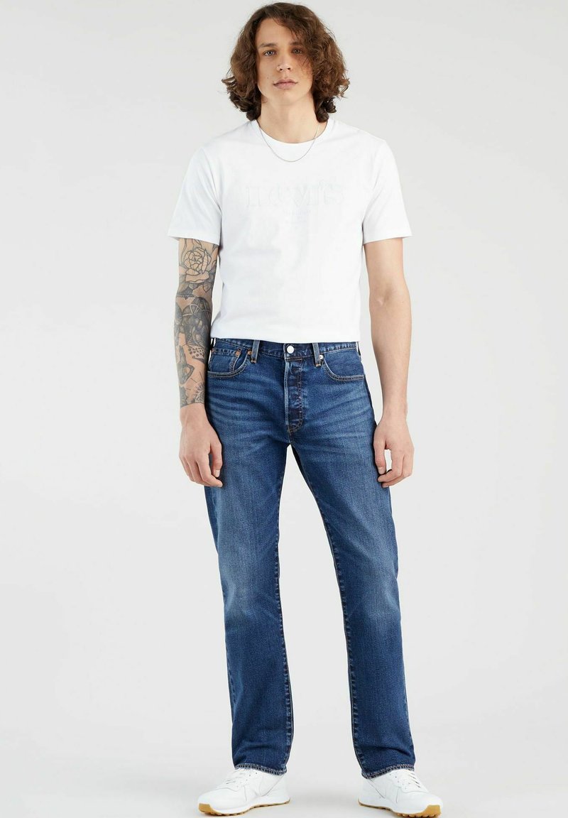 Blauwe spijkerbroek met een rechte snit, gecombineerd met een witte t-shirt met ronde hals voorzien van een subtiel logo. Witte sneakers maken de look compleet.