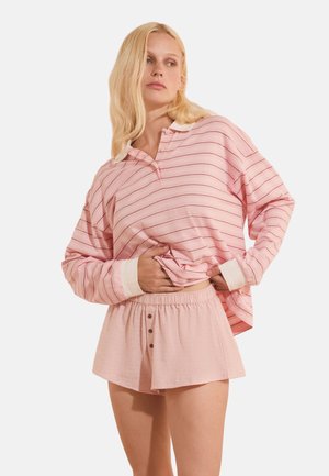 Donna bionda che indossa una camicia rosa a righe larga a maniche lunghe e pantaloncini rosa chiaro con bottoni, alzando leggermente la camicia, guardando di lato.