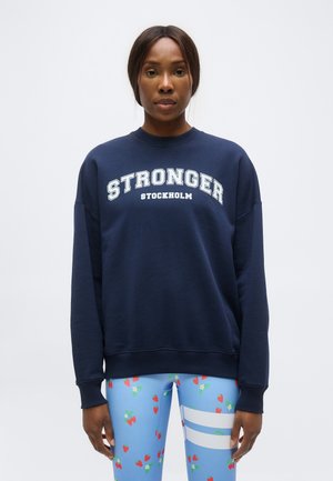 Femme portant un sweat-shirt bleu marine avec le texte « STRONGER STOCKHOLM » et des leggings bleu clair avec des motifs de fraises et de rayures blanches.