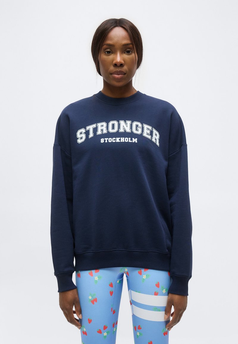 Femme portant un sweat-shirt bleu marine avec le texte « STRONGER STOCKHOLM » et des leggings bleu clair avec des motifs de fraises et de rayures blanches.