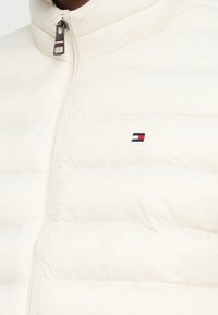 Valge turske vest kõrge kraega, horisontaalse õmblusega ja tõmblukuga. Omab väikest tikitud logo sinise, punase ja valgega.