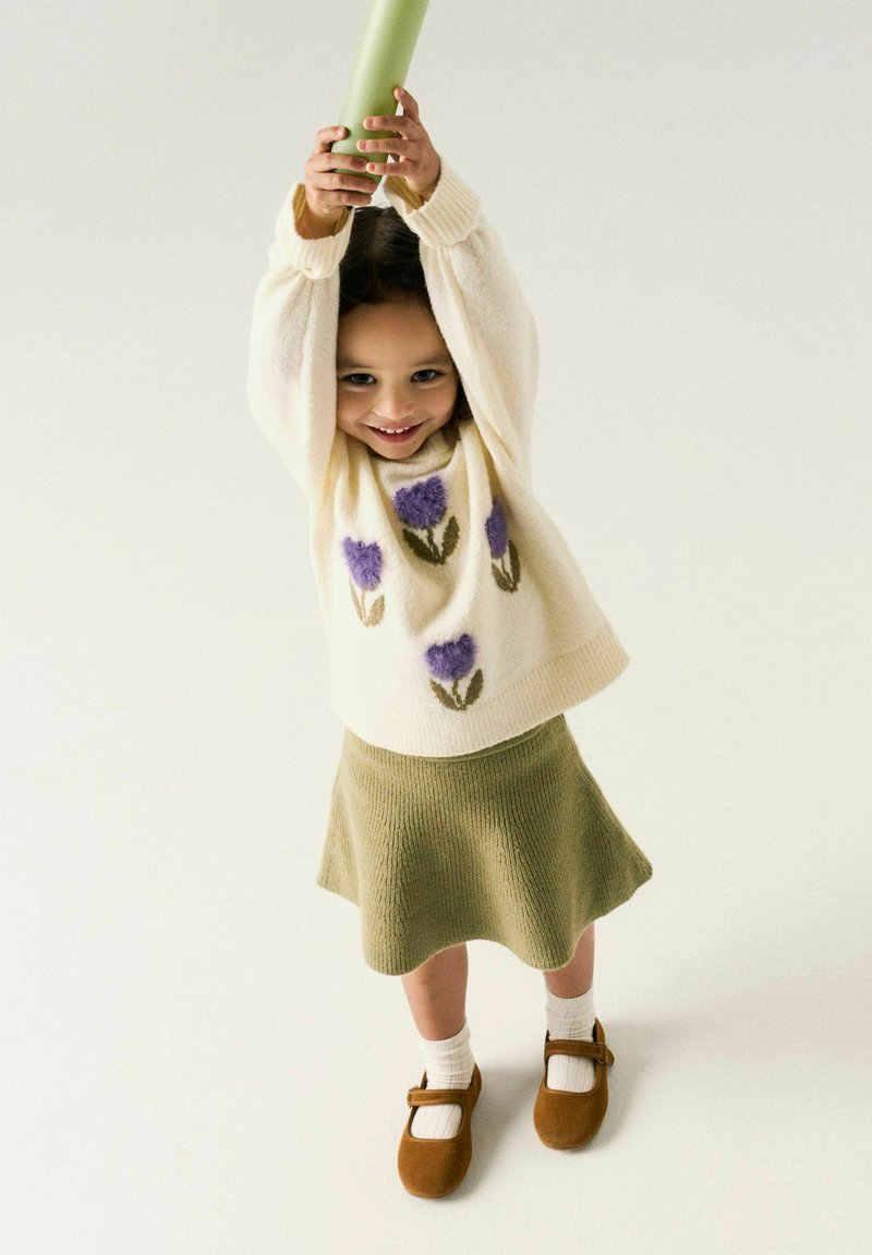 Jeune fille souriante, portant un pull crème avec des fleurs violettes, une jupe verte et des chaussures marron, tenant un objet vert au-dessus de sa tête.