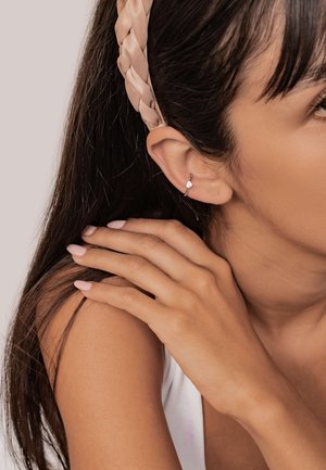 Silver hoop earring with a small diamond accent. Light brown braided headband. Close-up of ear, hand resting on shoulder, smooth skin texture.