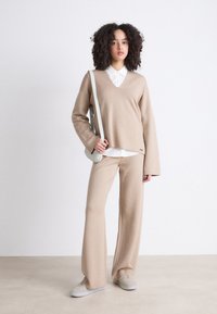 Maglione beige lavorato a maglia con scollo a V, abbinato a pantaloni larghi coordinati. Camicia bianca con colletto sotto; porta una borsa a tracolla grigia chiara.