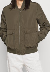 Olivově zelená bomber bunda se zapínáním na zip, žebrovanými manžetami a spodním lemem a dvěma předními kapsami. Hladká textura s minimálním zdobením.