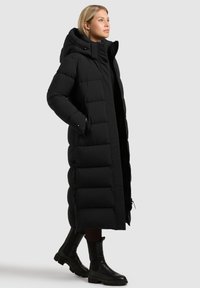 Manteau matelassé long noir avec capuche, présentant un design matelassé, une fermeture éclair frontale et des poches latérales. Associé à des bottes noires montantes jusqu'aux genoux.