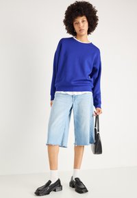 Felpa blu con maniche lunghe e collo alto, abbinata a pantaloncini di jeans blu chiaro. Il modello indossa calze bianche e mocassini neri, tenendo una borsa nera.