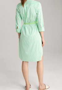 Sukienka koszulowa w zielono-białe paski z rękawami o długości do łokcia, paskiem do wiązania w talii oraz zaokrąglonym dołem. Posiada mankiety zapinane na guziki i boczne rozcięcia.