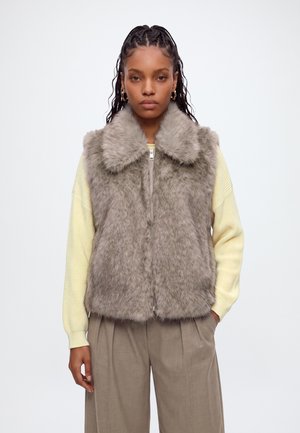 Femme portant un gilet sans manches en fausse fourrure grise sur un pull jaune clair et un pantalon beige, debout sur un fond blanc uni.