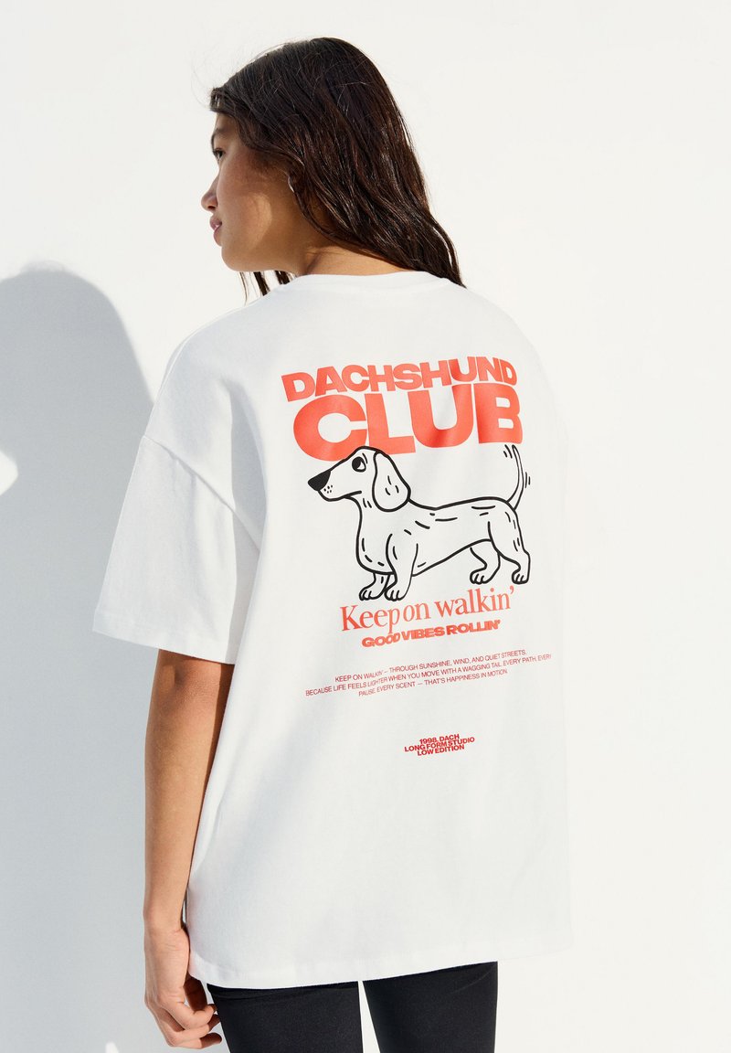 Femme aux longs cheveux foncés portant un t-shirt blanc oversize avec un texte rouge "Dachshund Club" et une illustration noire d'un teckel dans le dos.