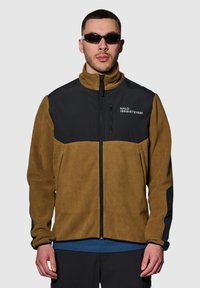 Bruine en zwarte fleecejas met een ritssluiting aan de voorkant, hoge kraag en zijzakken. Bevat een logo, contrasterende panelen en een gemêleerd uiterlijk.