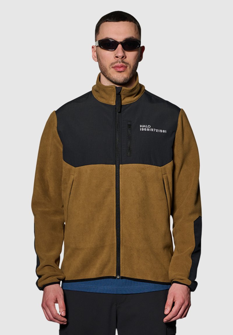 Bruine en zwarte fleecejas met een ritssluiting aan de voorkant, hoge kraag en zijzakken. Bevat een logo, contrasterende panelen en een gemêleerd uiterlijk.