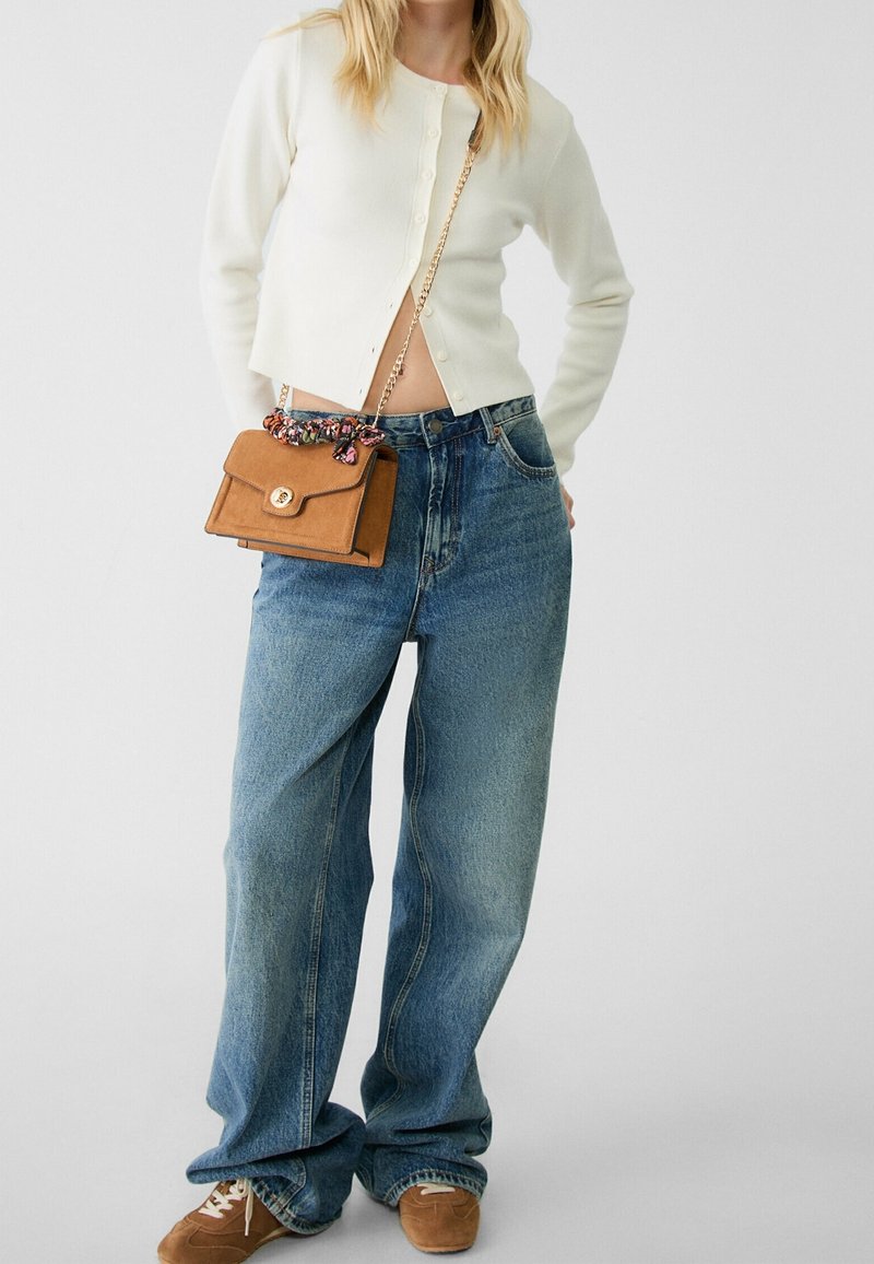 Femme portant un cardigan court blanc, un jean bleu ample, des baskets marron en daim, et portant un sac à bandoulière marron avec une chaîne et un détail foulard.