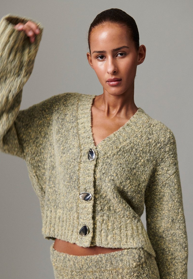 Cardigã verde texturizado em malha com decote em V, três grandes botões prateados e mangas largas. Design ajustado e cropped com bainha canelada.