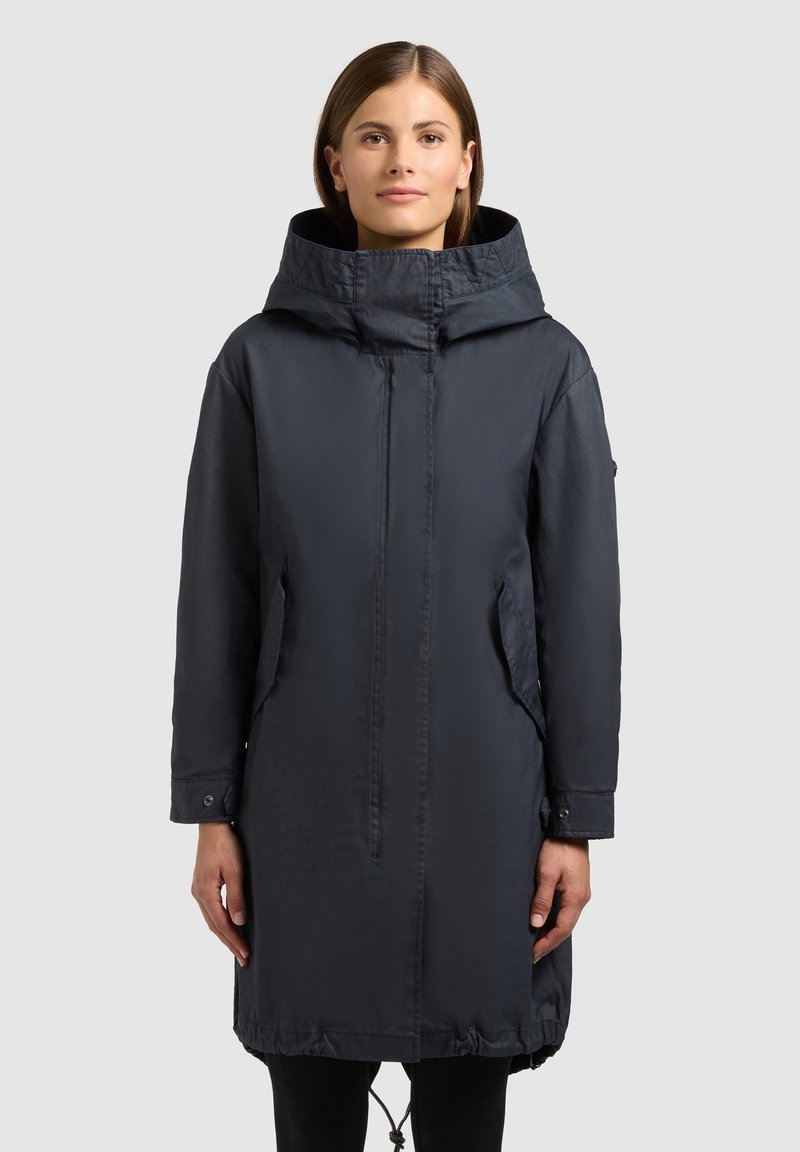 Kobieta nosząca długi, ciemnoniebieski płaszcz z kapturem, zapinany na zamek błyskawiczny, z przednimi kieszeniami, stojąca na jednolitym jasnoszarym tle.