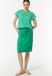 Green patterned top with short sleeves, paired with a matching green drawstring skirt. Model wears beige strappy sandals.