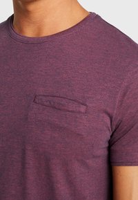 Camiseta de manga corta de color púrpura oscuro con escote redondeado y un pequeño bolsillo en el lado izquierdo. La tela tiene un aspecto texturizado.