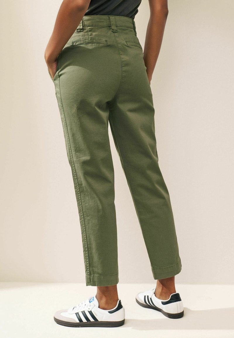 Personne portant un pantalon vert taille haute et des baskets blanches avec des bandes noires, debout devant un fond clair uni.