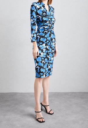 Mujer con un vestido ajustado de flores con patrones azules, blancos y negros, mangas largas y sandalias negras de tacón alto sobre un suelo gris.