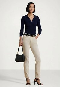 Mulher vestindo uma blusa de manga longa azul-marinho, calças bege com cinto preto, sandálias de salto alto pretas, segurando uma bolsa preta, em pé contra um fundo simples.