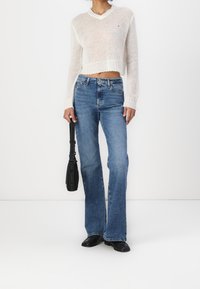 Suéter de punto blanco, recortado, con escote en V; jeans azules acampanados; bolso negro con correa; calzado negro; fondo liso.