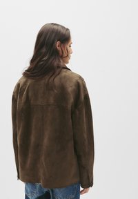 Hnědá semišová bunda s volným střihem, která má šev na zadním díle a knoflíkové manžety. Nosená přes modré denimové kalhoty, minimalistický design.