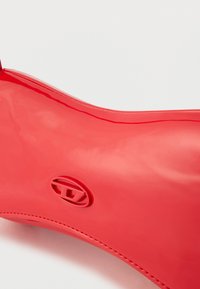 Spīdīga sarkana gumijas lietus zābaki ar noapaļotu purngalu un reljefu logotipu sānos, attēlots uz baltas fonas.