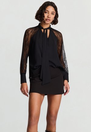 Donna che indossa una camicetta nera con maniche in pizzo e un fiocco sul davanti, abbinata a una minigonna nera, su uno sfondo chiaro e uniforme.