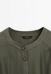 Olivenfarvet skjorte med rund halsudskæring, der har to knapper foran og tekstureret stof. Mærket viser "Massimo Dutti, Made in China."