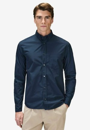 Ung mand iført en knappet marineblå skjorte med lange ærmer og brystlomme samt beige bukser, stående mod en ensfarvet hvid baggrund.