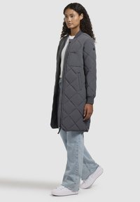 Geverfde grijze pufferjas met een rits aan de voorkant, zijzakken en ribgebreide manchetten; gestyled met lichtblauwe jeans en witte sneakers.