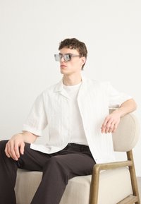 Witte shirt met korte mouwen en een gestructureerd ontwerp, gedragen over een wit T-shirt; donkere broek; glazen zonnebril met hoekige frames.