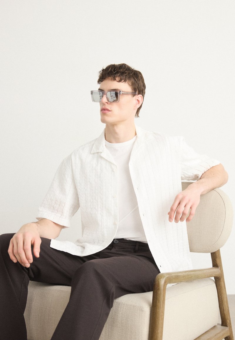 Witte shirt met korte mouwen en een gestructureerd ontwerp, gedragen over een wit T-shirt; donkere broek; glazen zonnebril met hoekige frames.