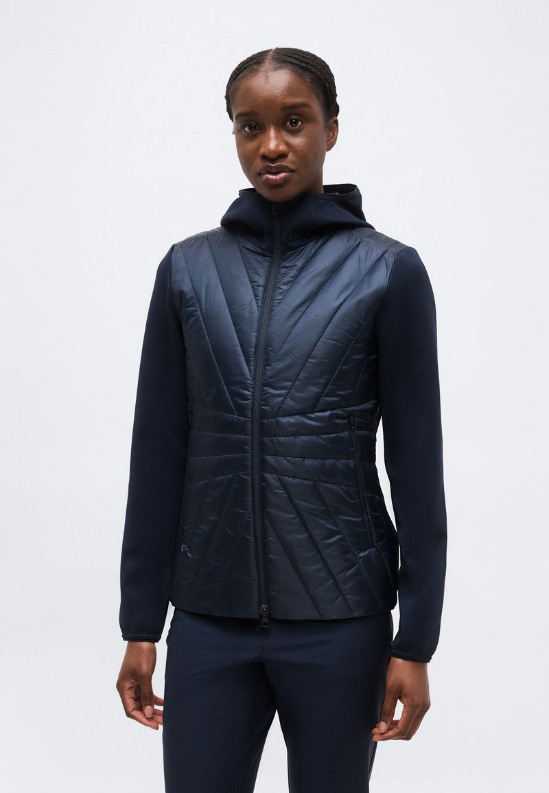 Femeie purtând o geacă matlasată bleumarin cu fermoar și glugă, și pantaloni asortați bleumarin, stând în fața unui fundal alb simplu.